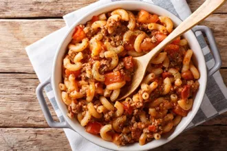 Overhead view of a serving pan of chili tomato macaroni. The pan rests on clothe squares on a wooden surface, with a wooden spoon in the chili.