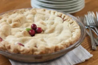 A baking dish containing an apple cranberry pie rests on a cloth. In the background are a stack of plates and loose pile of forks.