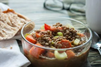 Close up view of a bowl of lentil stew next to a cloth, spoon, and piece of bread.