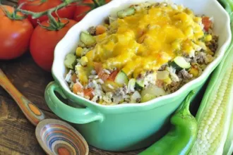 Overhead view of a baking dish of enchilada rice. A serving spoon, fresh tomatoes, an ear of corn, and an Anaheim paper.