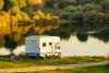 A recreational vehicle parked next to a pond or small lake with a solar panel behind it in a natural setting around sunset.